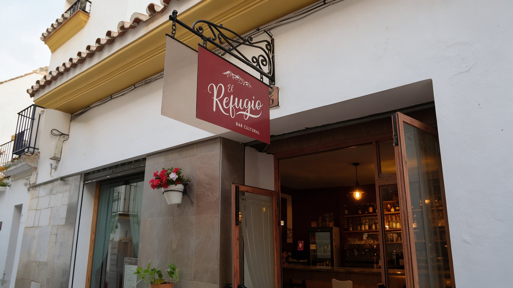 Rótulo granate colgante con el nombre El Refugio en letra blanca manuscrita y BAR CULTURAL debajo, sobre la puerta de un bar acogedor con geranios rojos